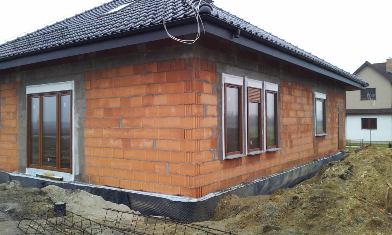 Surowy stan budynku w trakcie budowy z zamontowanymi oknami z brązowymi ramami i częściowo opuszczonymi roletami zewnętrznymi, widoczne pustaki ceramiczne i czarna folia fundamentowa.