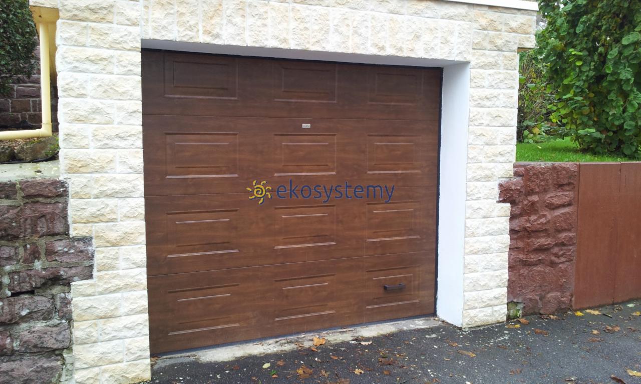 Brązowa segmentowa brama garażowa z widocznym logo firmy, zamontowana w jasnym, kamiennym murze.