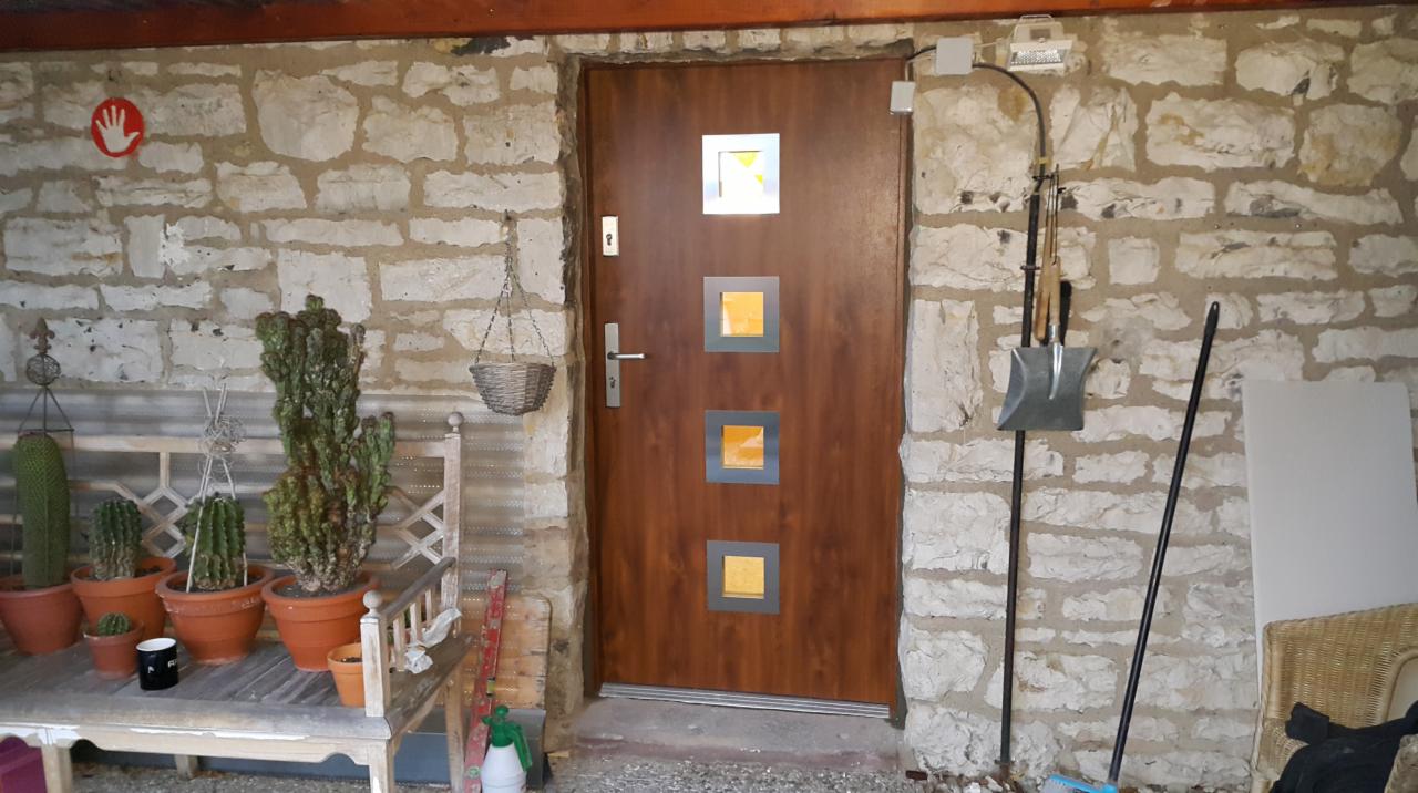 Brązowe drzwi wejściowe z trzema kwadratowymi okienkami z żółtym szkłem, osadzone w kamiennej ścianie, obok ławka z kaktusami w doniczkach i wiszący koszyk.