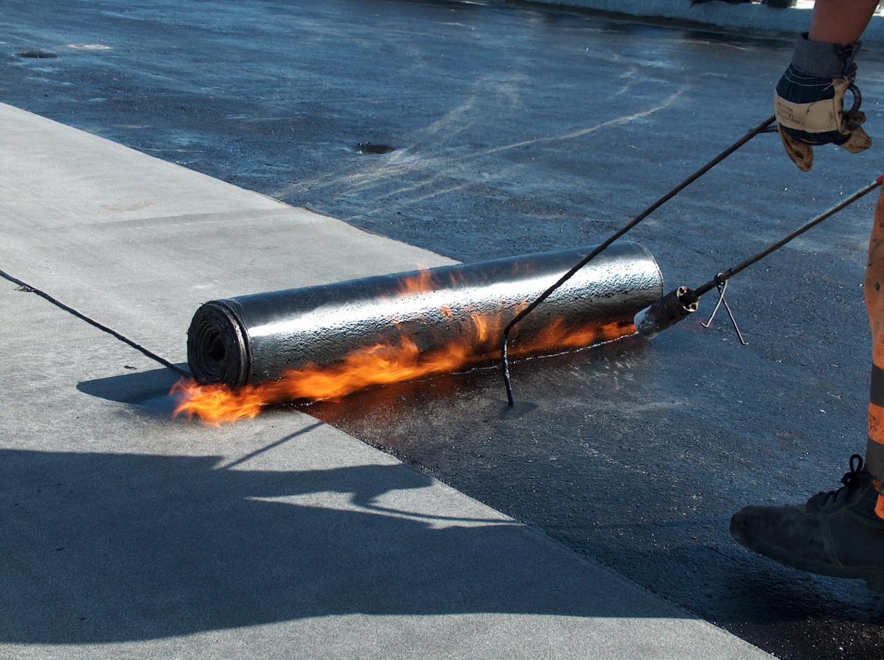 Układanie papy termozgrzewalnej na dachu płaskim z użyciem palnika, widoczne płomienie i narzędzia dekarskie.