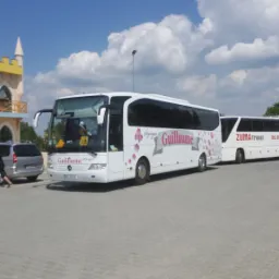 Dwa białe autokary turystyczne zaparkowane na brukowanym placu, z budynkiem w stylu zamku w tle, błękitne niebo z chmurami.