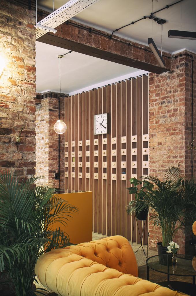 Wnętrze lokalu usługowego z elementami industrialnymi: ściana z czerwonej cegły, drewniana ścianka działowa z zegarem, żółta sofa Chesterfield i rośliny doniczkowe.