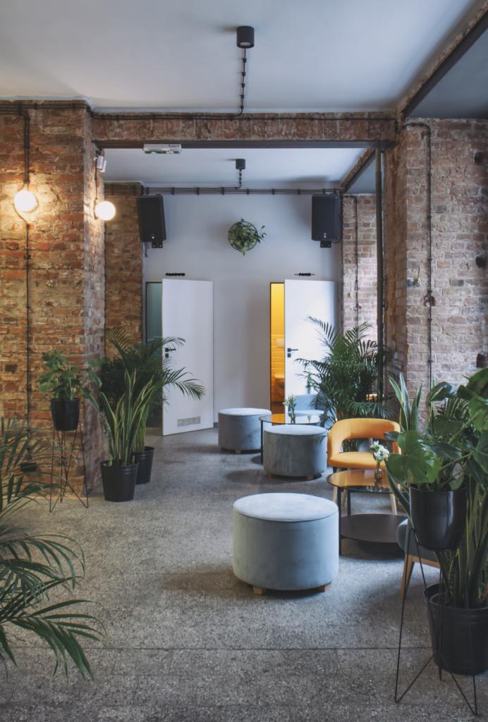 Wnętrze lokalu usługowego z surowymi ceglanymi ścianami, szarymi pufami, roślinami doniczkowymi i żółtym krzesłem, widoczne drzwi do toalet z podświetleniem.