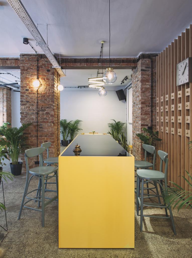 Wnętrze lokalu usługowego z żółtą ladą barową, szarymi krzesłami barowymi, ceglanymi kolumnami, roślinami doniczkowymi i zegarem na drewnianej ścianie z listew.