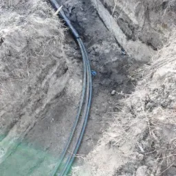 Wąski wykop w ziemi z ułożonymi rurami, widoczne nierówne ściany wykopu i fragmenty korzeni.