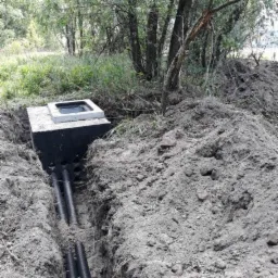 Betonowa studzienka z pokrywą w wykopie, widoczne rury doprowadzające, ziemia usypana w kopiec, w tle drzewa.