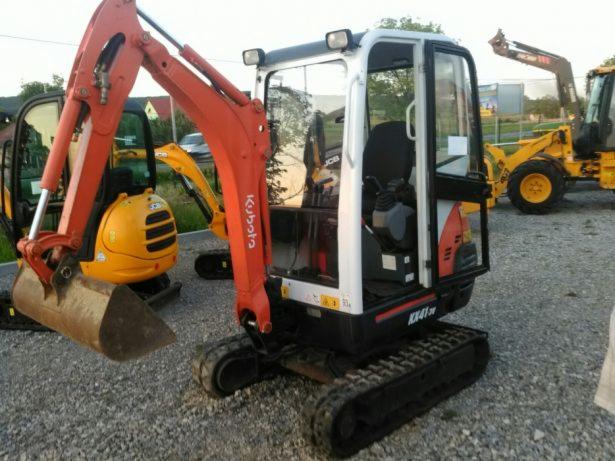 Pomarańczowa mini koparka Kubota KX41-3V z łyżką z przodu, widoczne gąsienice, kabina z szybami i żółta koparka JCB w tle na żwirowym podłożu.