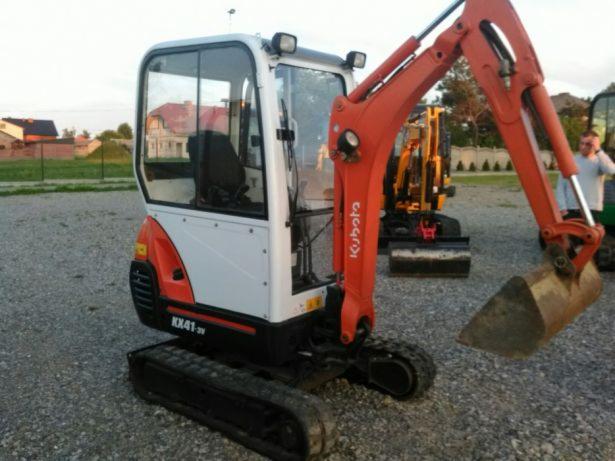 Kompaktowa mini koparka Kubota KX41-3V z łyżką, widoczna kabina operatora, ramię koparki w kolorze pomarańczowym, w tle inne maszyny budowlane i zabudowa mieszkalna.