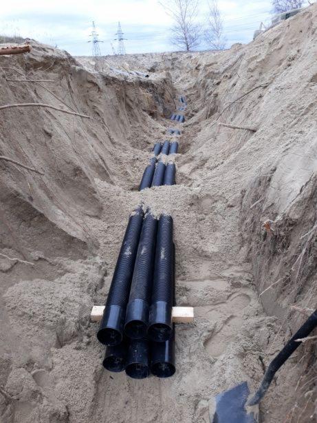Głęboki wykop w piaszczystej ziemi w Lubajnach, z ułożonymi czarnymi rurami na drewnianych podporach, widoczne ślady pracy ziemnej i linia wysokiego napięcia w tle.
