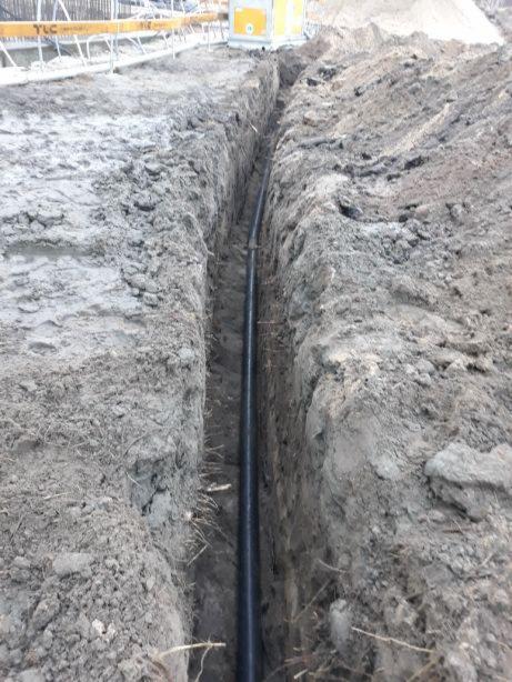 Wąski wykop w ziemi z widoczną czarną rurą na dnie, obok żółte barierki zabezpieczające teren budowy.