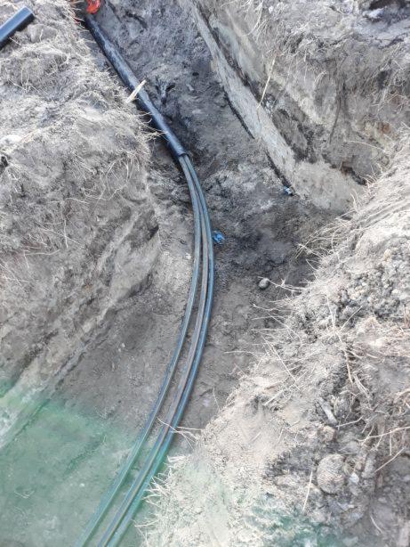 Wąski wykop w ziemi z ułożonymi rurami, widoczne nierówne ściany wykopu i fragmenty korzeni.