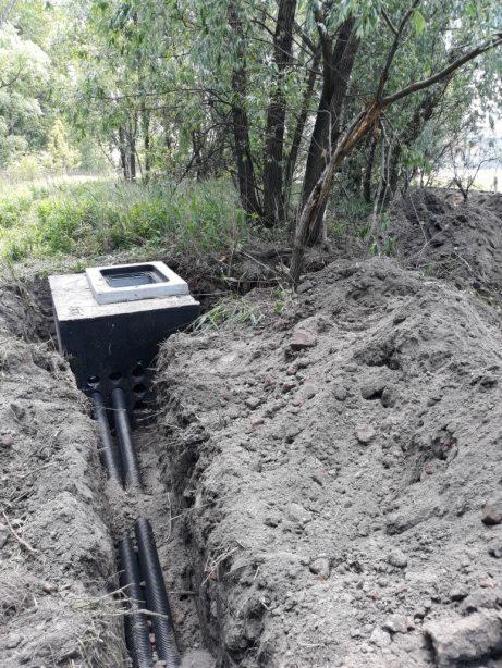 Betonowa studzienka z pokrywą w wykopie, widoczne rury doprowadzające, ziemia usypana w kopiec, w tle drzewa.