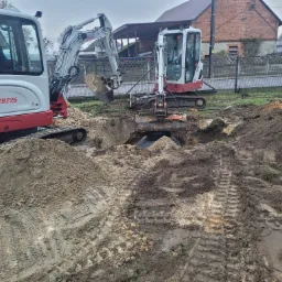 Dwa mini-koparki TB225, jedna z operatorem, pracują w wykopie na działce z ogrodzeniem, w tle dom z czerwonej cegły i pochmurne niebo.