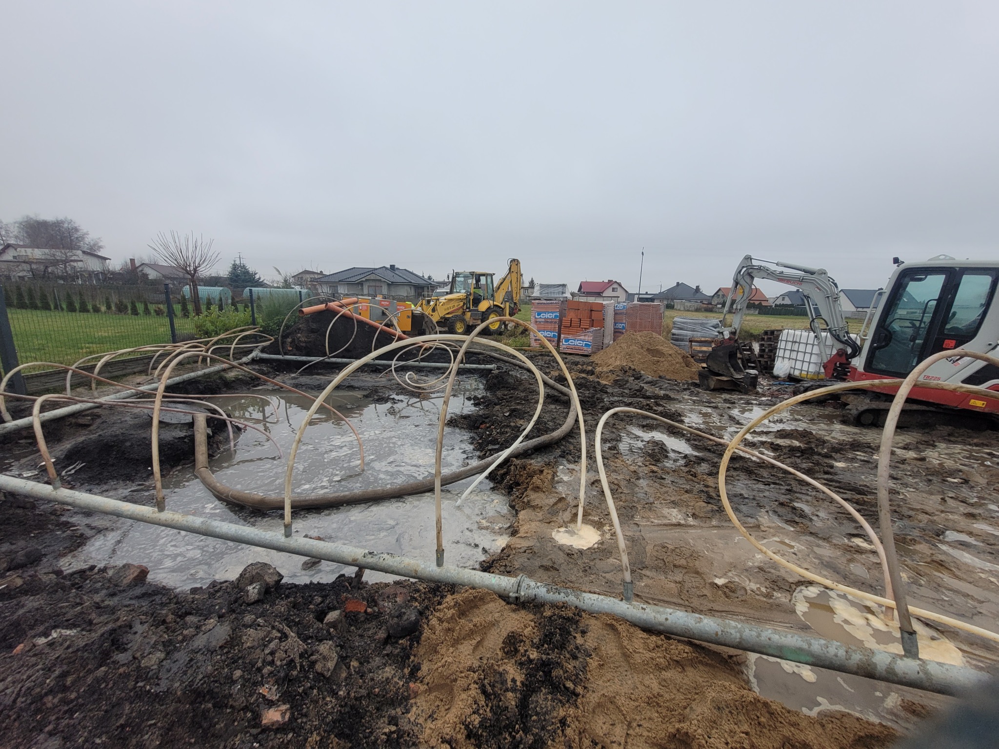 Przygotowanie terenu pod budowę: widoczne rury ułożone w wykopie wypełnionym wodą, w tle koparki i palety z cegłami. Pochmurne niebo.