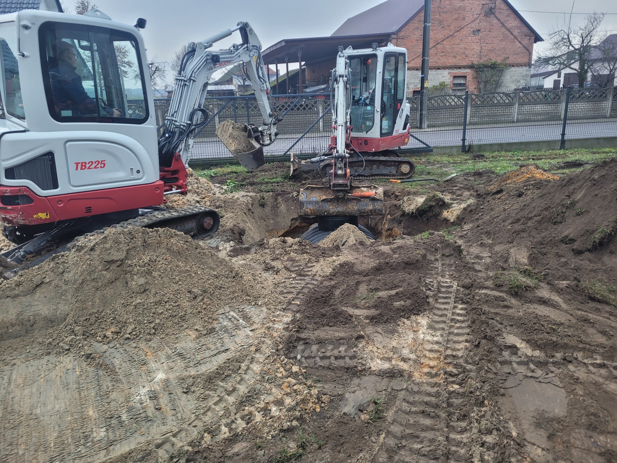 Dwa mini-koparki TB225, jedna z operatorem, pracują w wykopie na działce z ogrodzeniem, w tle dom z czerwonej cegły i pochmurne niebo.
