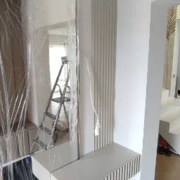 Montaż toaletki z lustrem w przedpokoju, lustro zabezpieczone folią ochronną, widoczna drabina w odbiciu, dekoracyjne panele ścienne w pionowe żłobienia.