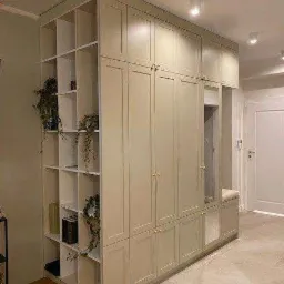 Duża, beżowa szafa wnękowa z otwartymi półkami po lewej stronie, dekorowana roślinami i książkami, z siedziskiem i lustrem po prawej stronie, w jasnym wnętrzu.