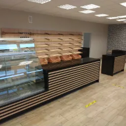 Wnętrze piekarni: przeszklona lada chłodnicza z poziomymi pasami, drewniane półki z wiklinowymi koszykami, wzorzysta tapeta w tle.