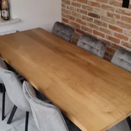 Drewniany stół jadalny z czarnymi, metalowymi nogami w stylu industrialnym, otoczony szarymi krzesłami na tle ściany z czerwonej cegły.