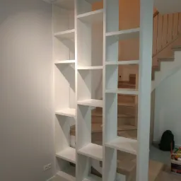 Biały regał dzielący przestrzeń, z widocznymi schodami w tle, drewniana podłoga, minimalistyczny design.