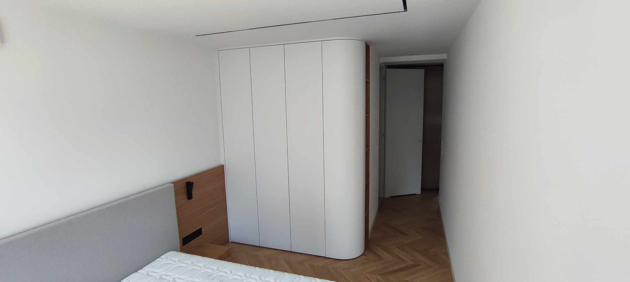 Nowoczesna szafa wnękowa z zaokrąglonym bokiem w minimalistycznej sypialni z parkietem w jodełkę i wejściem do garderoby.