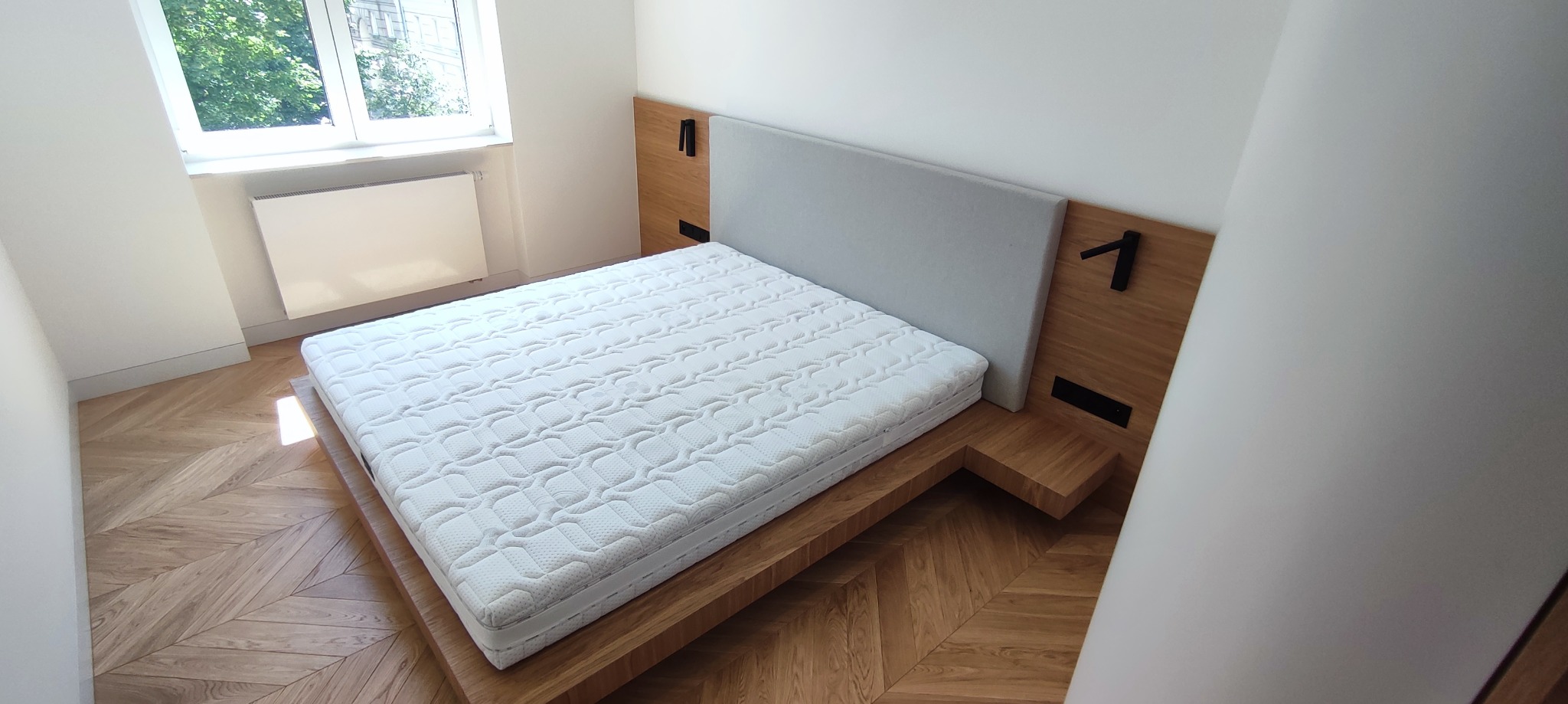 Nowoczesne łóżko na drewnianej ramie z szarym zagłówkiem, z czarnymi elementami dekoracyjnymi, na parkiecie ułożonym we wzór jodełki.