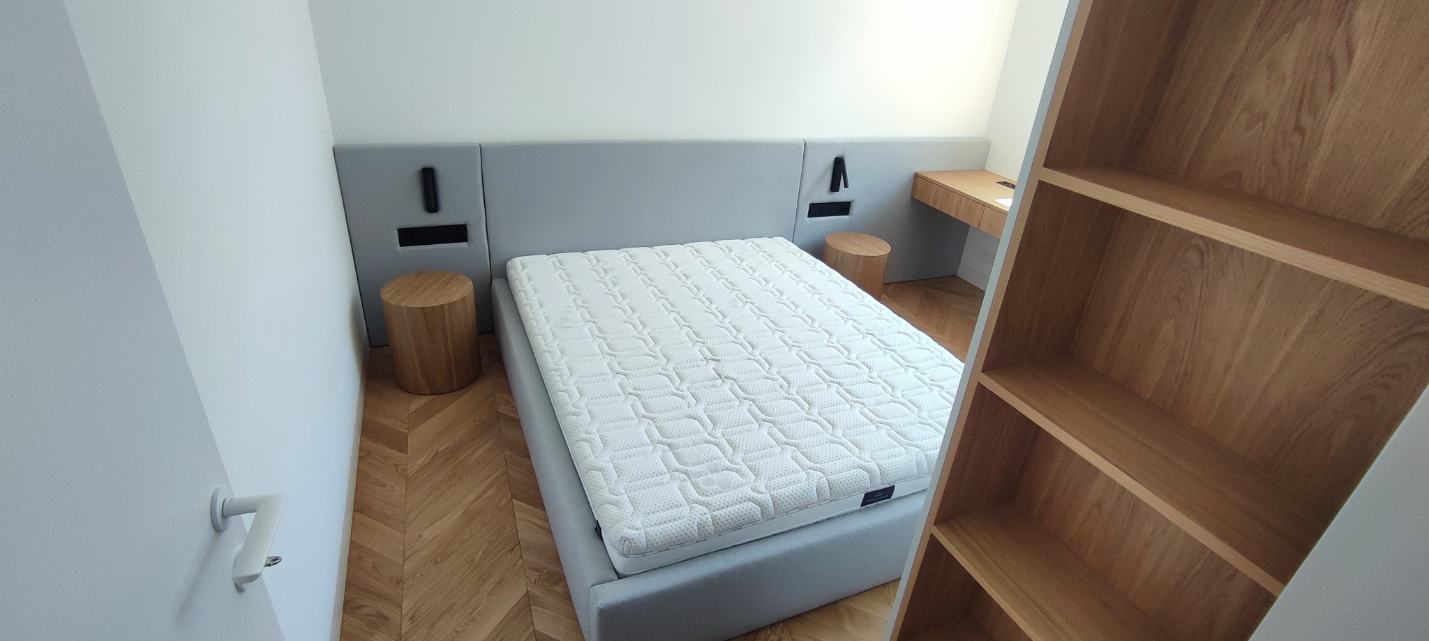 Nowoczesna sypialnia z łóżkiem kontynentalnym, szarym zagłówkiem z wbudowanymi gniazdkami, drewnianymi szafkami nocnymi w kształcie pniaków, biurkiem i regałem z jasnego drewna na parkiecie w jodełkę.