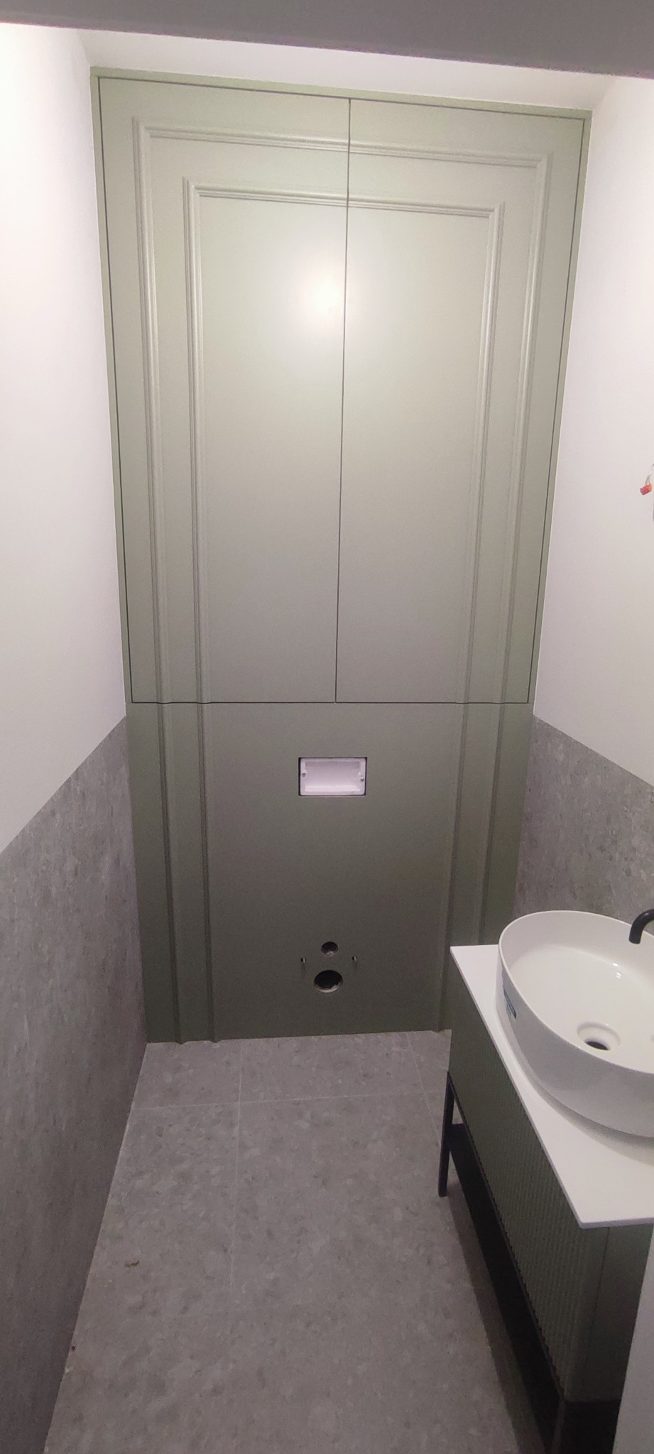 Zabudowa toalety podtynkowej z szafkami w kolorze oliwkowej zieleni, umywalka nablatowa na szafce, szara płytka na podłodze i ścianach.