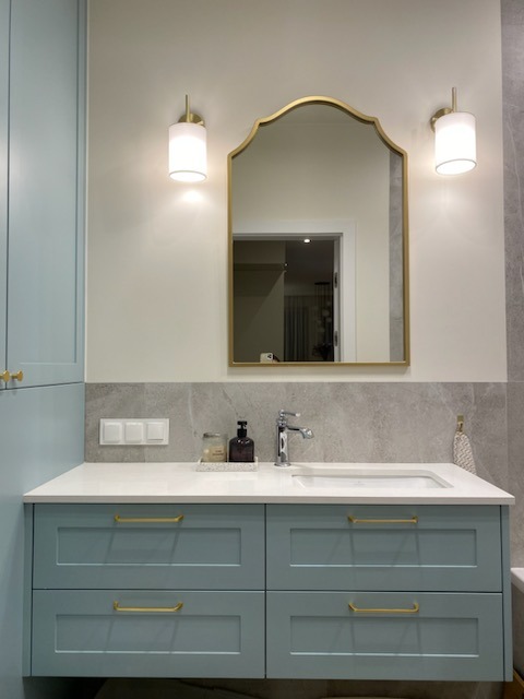 Niebieska szafka łazienkowa na wymiar z białym blatem i złotymi uchwytami, nad którą wisi lustro w złotej ramie i dwie lampki ścienne.