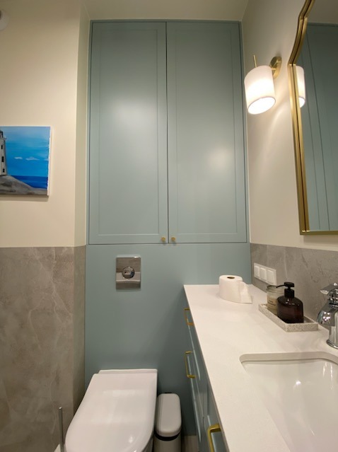 Jasna łazienka z zabudową meblową w pastelowym odcieniu, wisząca toaleta z chromowanym przyciskiem spłukiwania, biały blat z dozownikiem mydła w odcieniach brązu, lustro w złotej ramie i obraz...