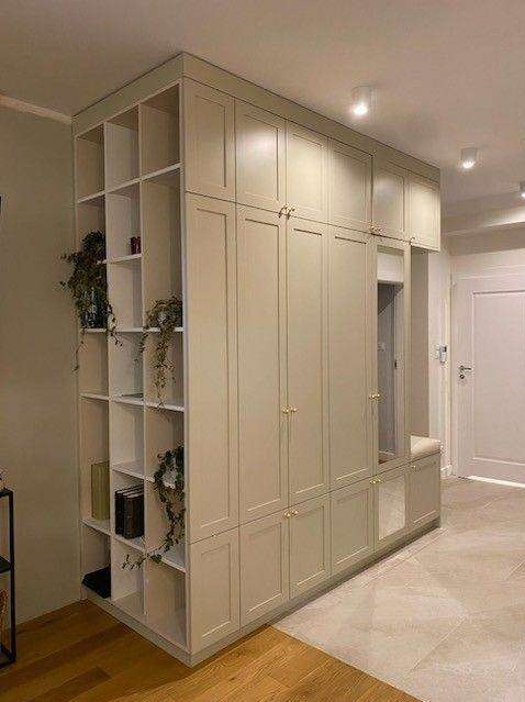 Duża, beżowa szafa wnękowa z otwartymi półkami po lewej stronie, dekorowana roślinami i książkami, z siedziskiem i lustrem po prawej stronie, w jasnym wnętrzu.