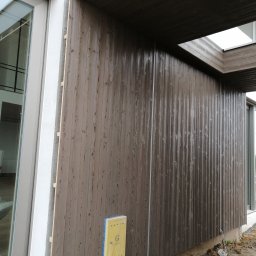 Tarasy Drewniane i Kompozytowe Arkadiusz Młynarski - Montaż pionowej, brązowej boazerii na elewacji budynku z dużym oknem; widoczny poziomica oparta o elewację i materiały budowlane wewnątrz budynku.