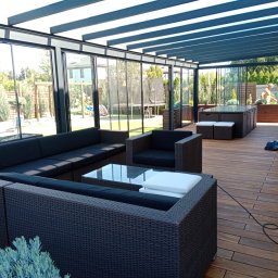 Tarasy Drewniane i Kompozytowe Arkadiusz Młynarski - Zadaszony taras z kompozytową deską, wyposażony w meble ogrodowe z ciemnej plecionki oraz szklane ściany przesuwne, widok na ogród z trampoliną i osobą bez koszulki.