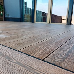 Tarasy Drewniane i Kompozytowe Arkadiusz Młynarski - Szczegółowe ujęcie desek tarasowych z wyraźnym rysunkiem słojów, ułożonych na zewnątrz budynku, z widocznym fragmentem ściany i okna, oraz zwiniętym wężem ogrodowym.