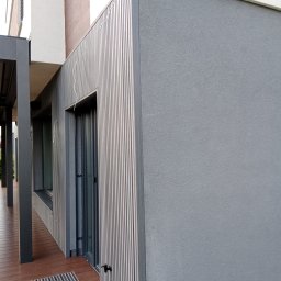 Tarasy Drewniane i Kompozytowe Arkadiusz Młynarski - Szara elewacja budynku z pionowymi panelami, brązowy taras z metalową kratką odpływową, widok z boku na fragment budynku.