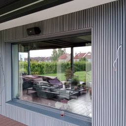 Tarasy Drewniane i Kompozytowe Arkadiusz Młynarski - Szara elewacja z pionowych paneli, z widocznym głośnikiem Bose i wystającym kablem, okno odbija taras z meblami ogrodowymi w Radomiu.