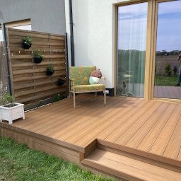 Tarasy Drewniane i Kompozytowe Arkadiusz Młynarski - Kompozytowy taras w odcieniach brązu, z niskim stopniem, otoczony zielonym trawnikiem, z drewnianym parawanem i wiszącymi doniczkami, krzesłem z kolorowymi poduszkami i widokiem na dom...