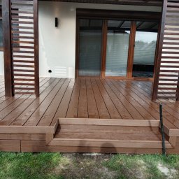 Tarasy Drewniane i Kompozytowe Arkadiusz Młynarski - Brązowy taras z desek z drewnianą pergolą i schodami, lekko mokry po deszczu, widok na biały budynek z oknami.