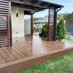 Tarasy Drewniane i Kompozytowe Arkadiusz Młynarski - Nowoczesny taras kompozytowy z drewnianą pergolą i zielonym trawnikiem po deszczu, widoczne krople wody na desce tarasowej.