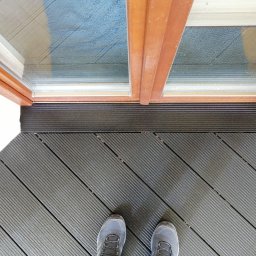 Tarasy Drewniane i Kompozytowe Arkadiusz Młynarski - Fragment tarasu z desek kompozytowych w Radomiu, widok z góry na szare buty na tle ułożonych desek, brązowa rama drzwi balkonowych z widocznym za szybą niebieskim materiałem.