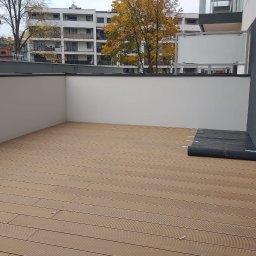 Tarasy Drewniane i Kompozytowe Arkadiusz Młynarski - Nowoczesny balkon z jasnobrązową podłogą z desek, widok na budynek mieszkalny z drzewami w jesiennych barwach, rolka czarnej folii leżąca przy ścianie.