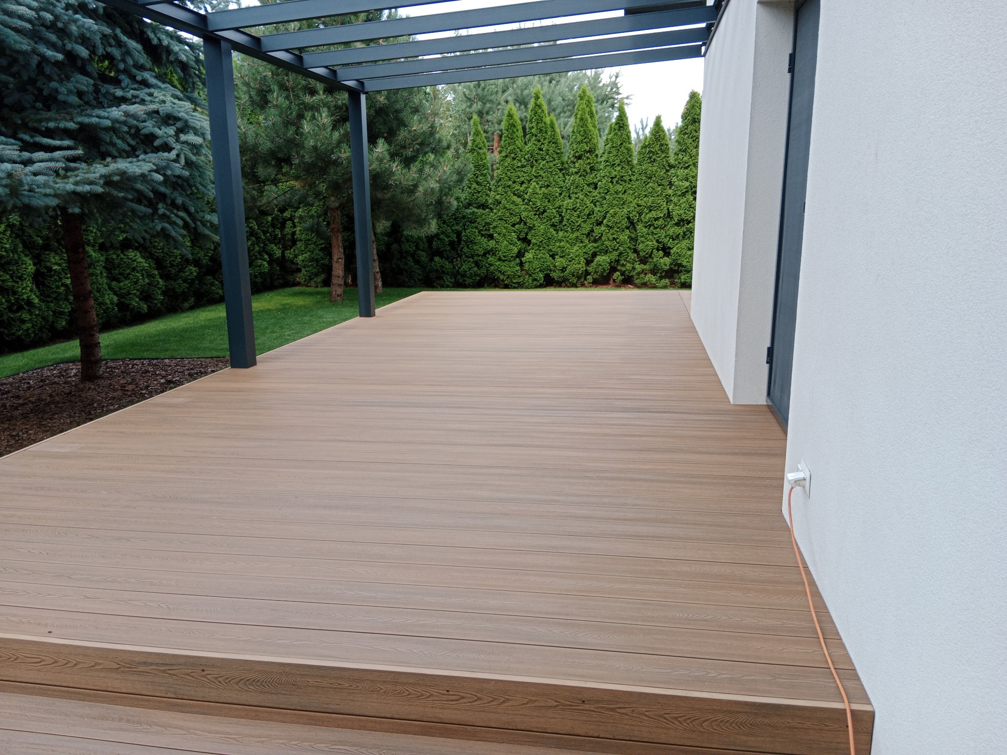 Jasnobrązowy taras z desek, zakończony schodkiem, z metalową pergolą, otoczony zielonymi drzewami iglastymi i trawnikiem, z widocznym gniazdkiem elektrycznym na ścianie budynku.