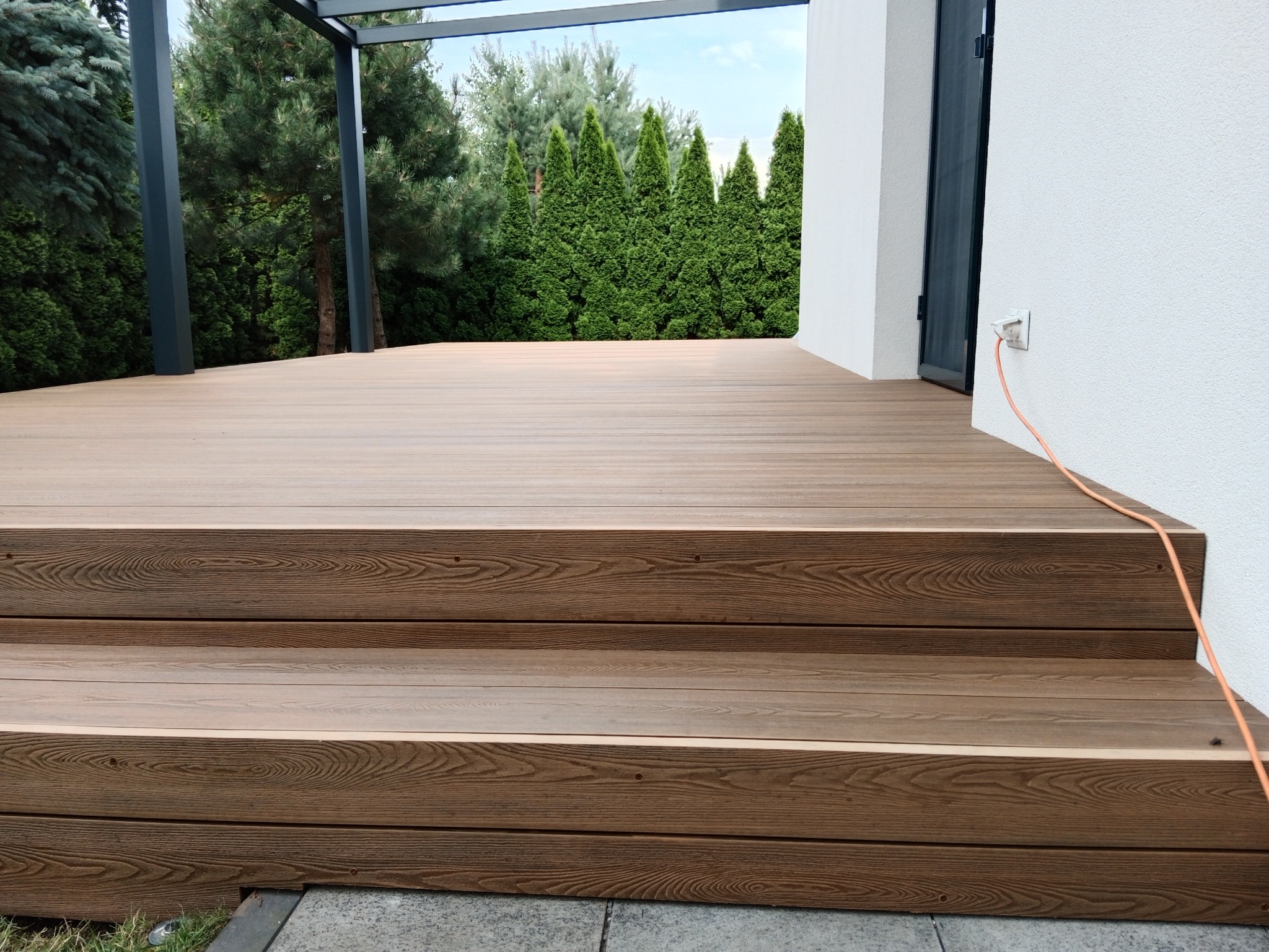 Kompozytowy taras z trzema stopniami, widok z dołu na zewnątrz, z zielonymi tujami w tle i kablem elektrycznym biegnącym wzdłuż ściany budynku.