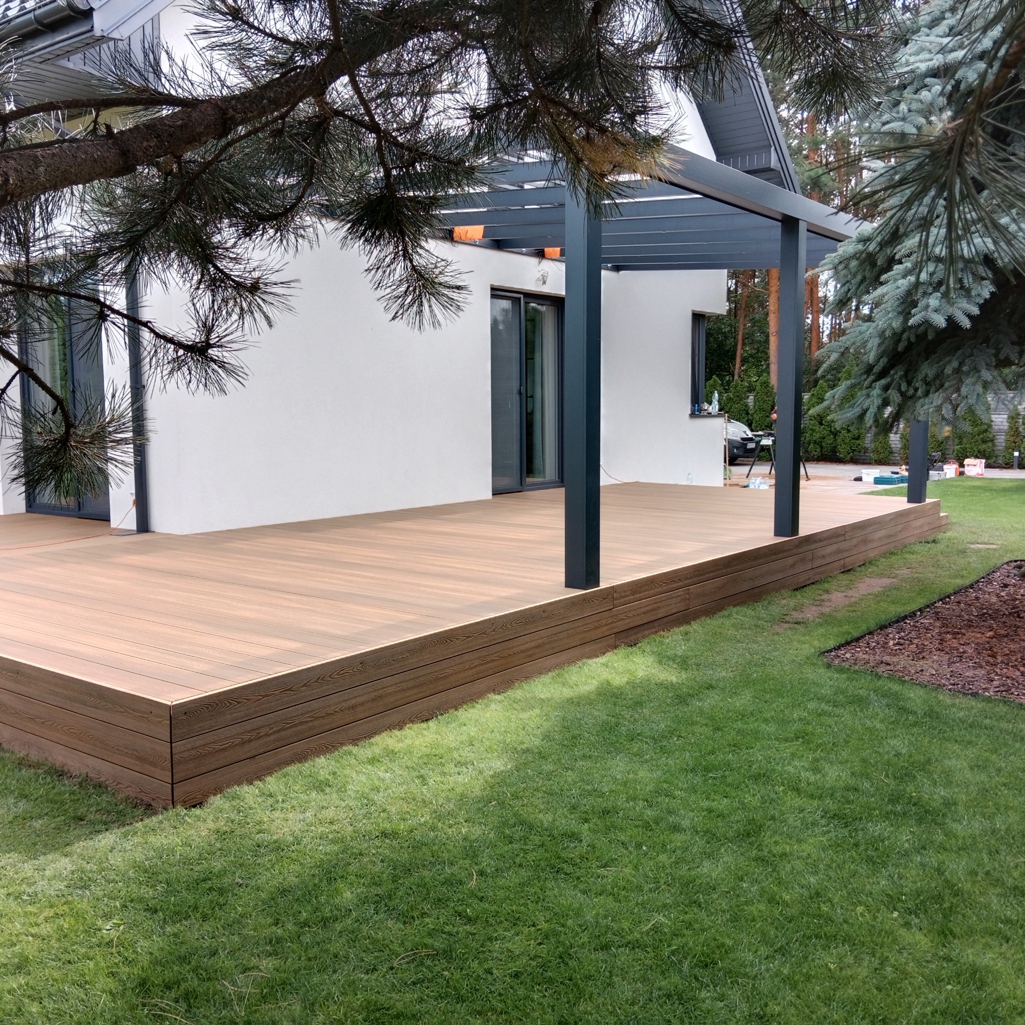 Nowoczesny taras z desek kompozytowych w Radomiu, zadaszony pergolą, przylegający do białego budynku z dużymi oknami, otoczony zielonym trawnikiem i drzewami iglastymi.