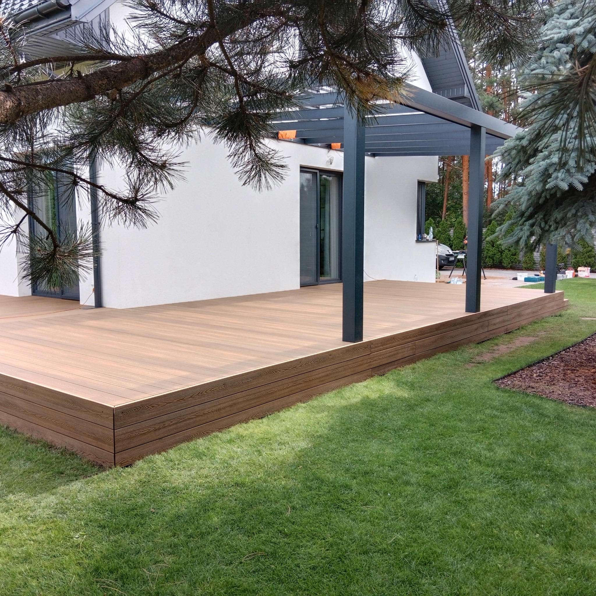 Nowoczesny taras z desek kompozytowych w kolorze jasnego drewna, zadaszony pergolą w kolorze grafitowym, przylegający do białego domu z dużymi oknami przesuwnymi, otoczony zielonym trawnikiem.