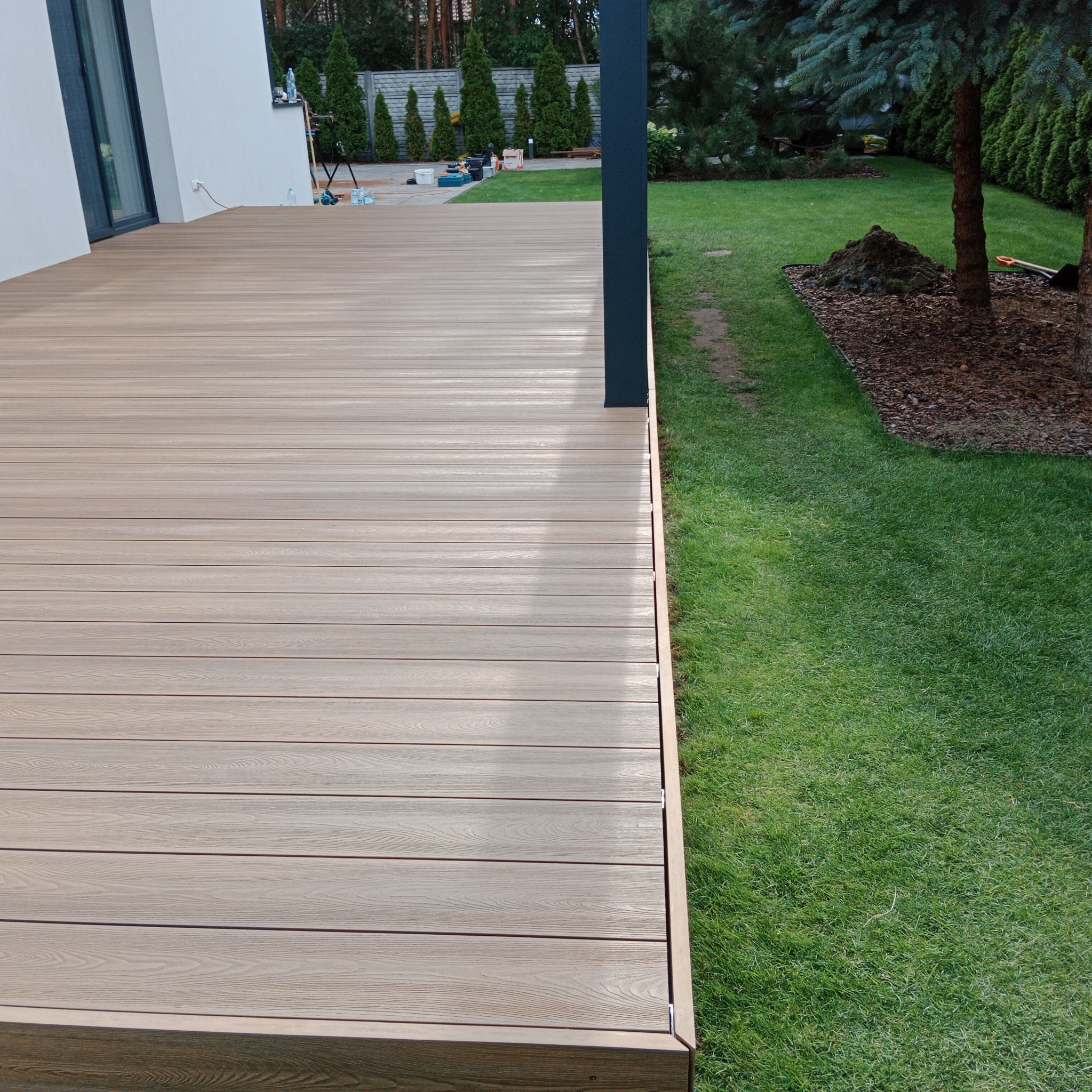 Nowoczesny taras z desek w kolorze jasnego drewna, z widocznym słupem konstrukcyjnym i trawnikiem w tle.