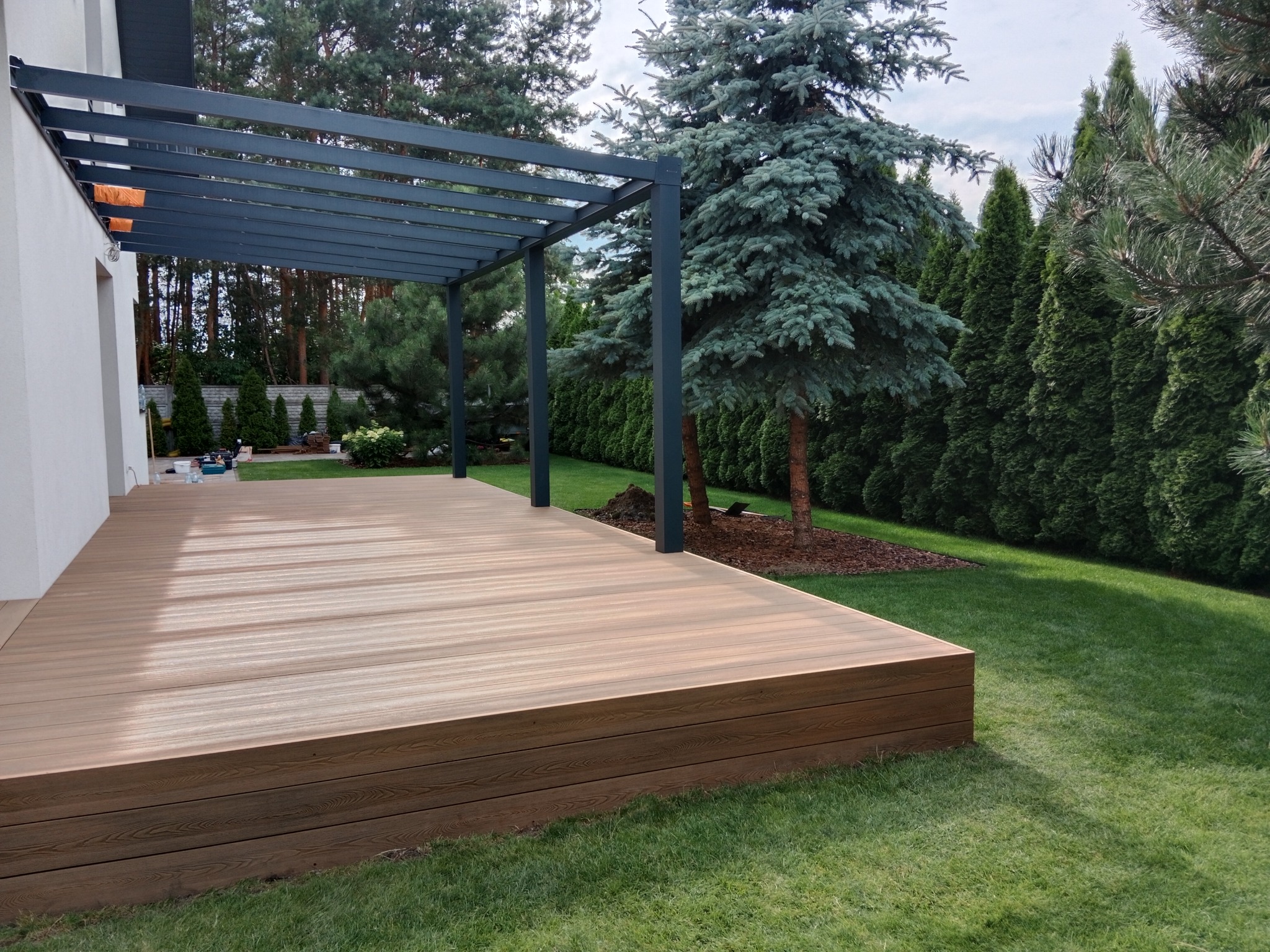 Jasna, kompozytowa deska tarasowa z metalową pergolą, otoczona zielonym trawnikiem i żywopłotem z iglaków, przylegająca do białej ściany budynku.