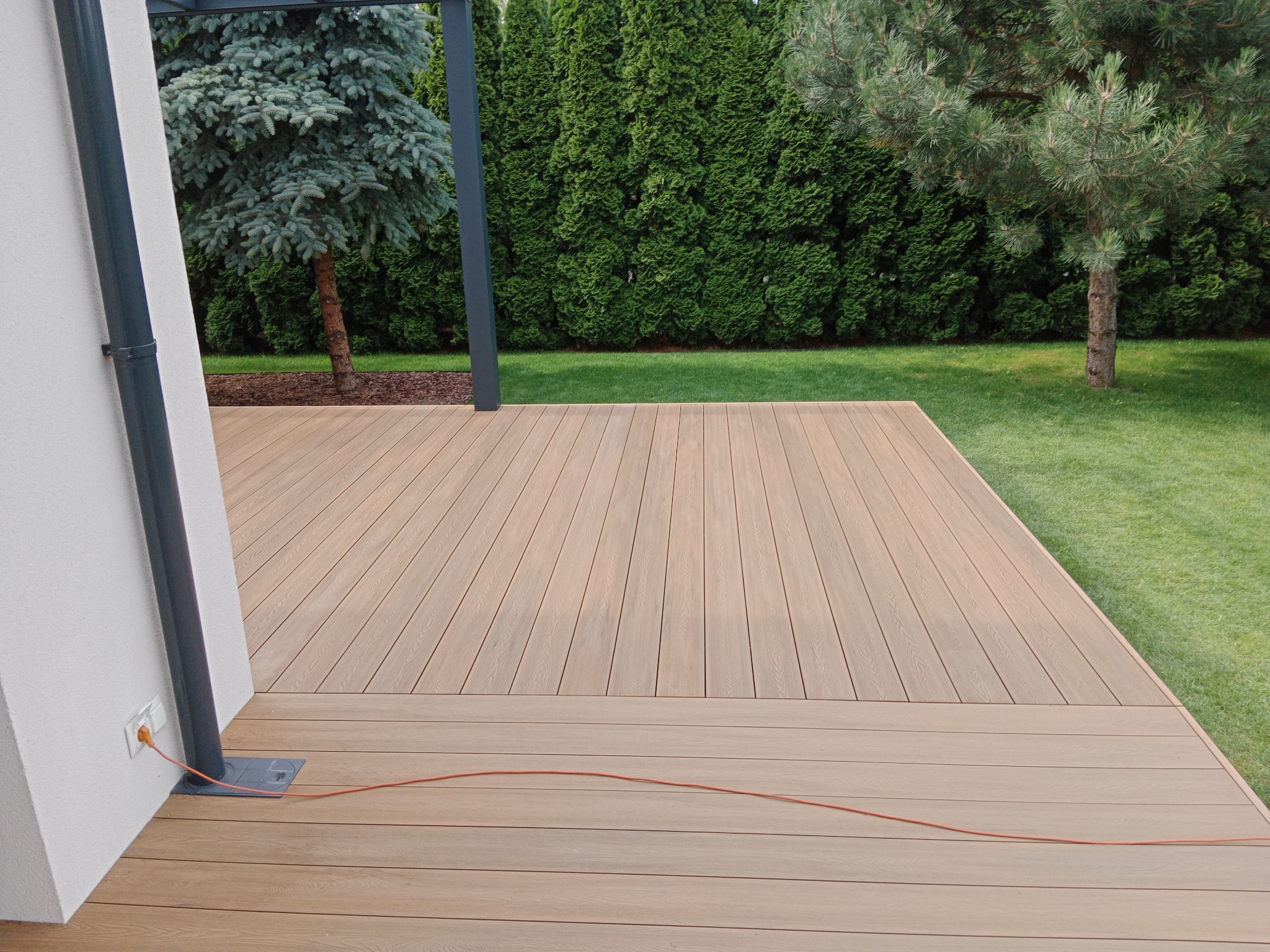 Jasna deska tarasowa z widocznym gniazdkiem elektrycznym na ścianie budynku, przewód w kolorze pomarańczowym leżący na desce, w tle trawnik i różne drzewa.