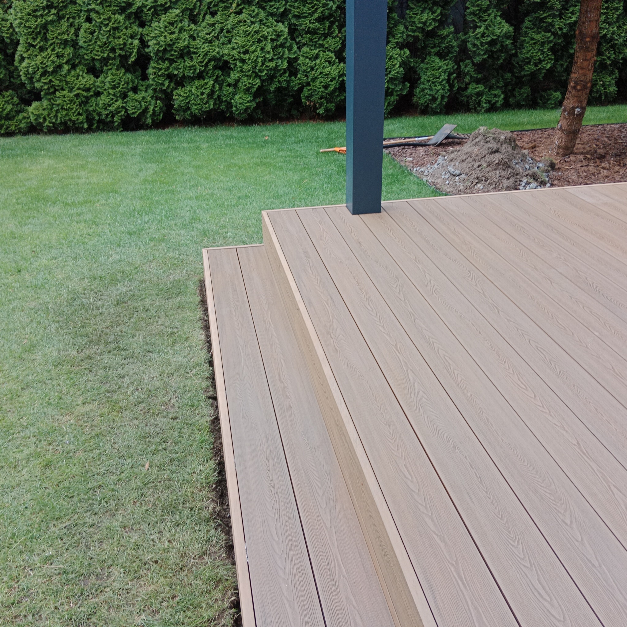 Nowoczesny taras z deski kompozytowej w odcieniach beżu, z dwoma stopniami, widok na zielony trawnik i żywopłot, czarny słupek podtrzymujący zadaszenie, narzędzia ogrodowe w tle.