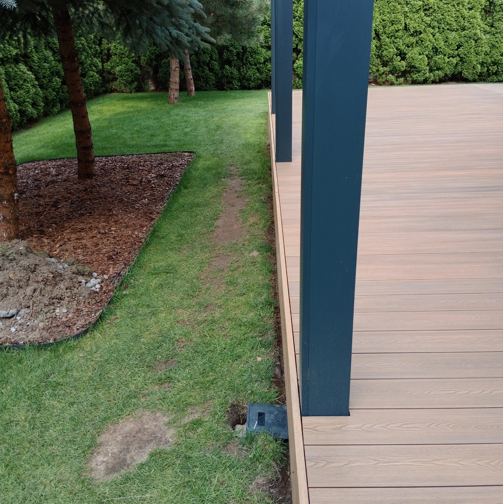 Nowoczesny taras z desek kompozytowych w odcieniach beżu, z ciemnymi słupkami konstrukcyjnymi, sąsiadujący z trawnikiem i rabatą z korą drzewną. Widoczna skrzynka instalacyjna w trawie przy...